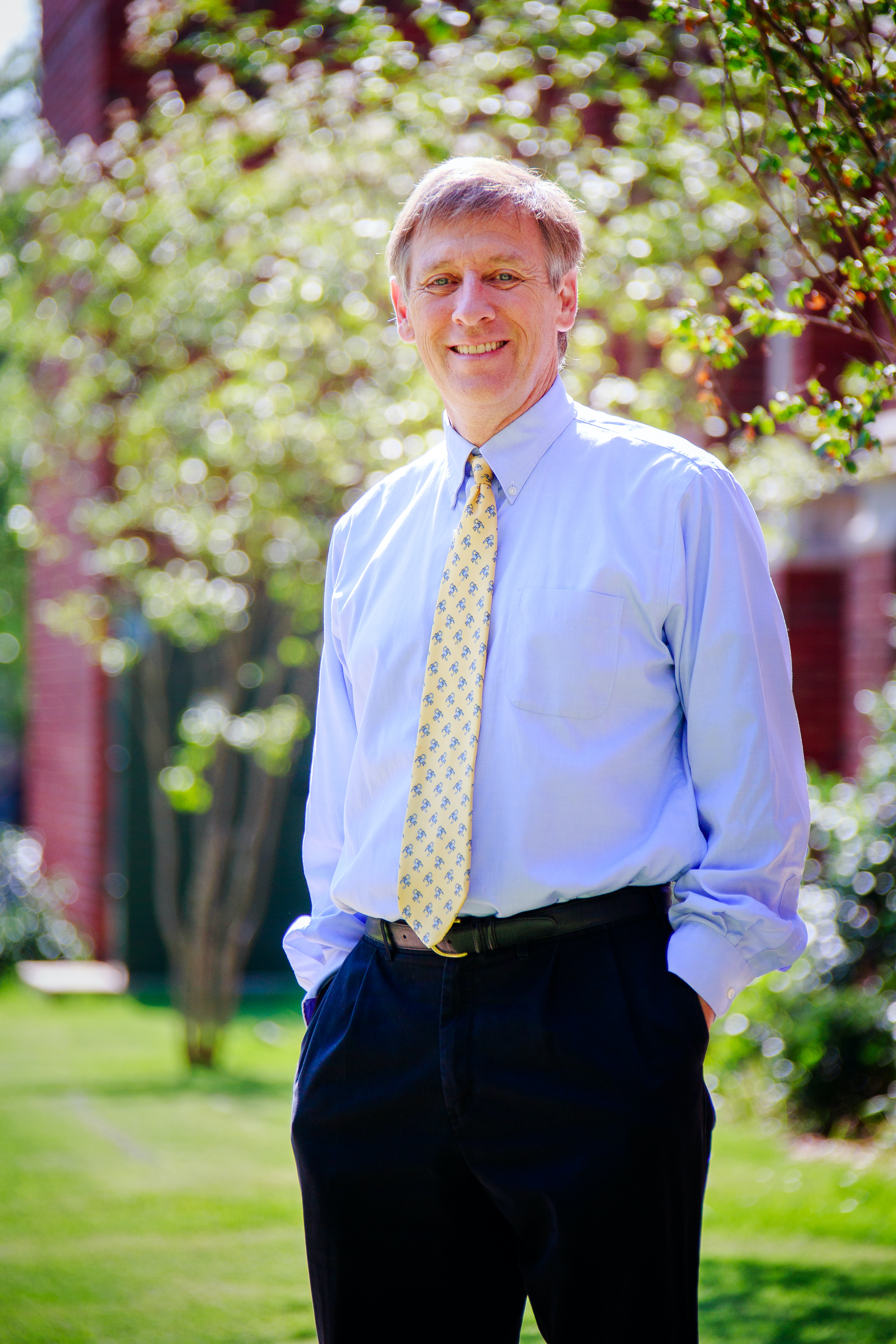 Doug Gaffin has short hair and is smiling. He is wearing a right blue, long-sleeved collared shirt, a yellow tie with blue pattern, and dark blue slacks. He stands outside with his hands in his pants pockets. In the background are blurred trees, a red brick building, and green grass.